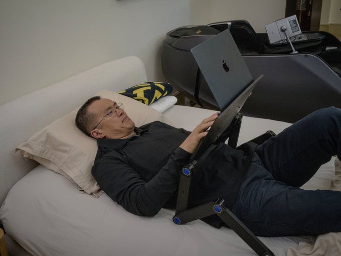 A man wearing glasses lies on a bed, working on a MacBook laptop placed on an adjustable stand over his lap.