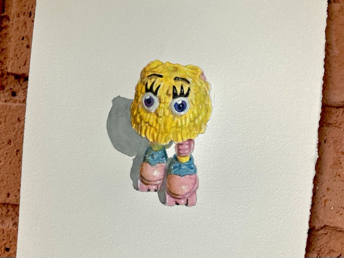 A small, handmade figurine of SpongeBob SquarePants with large purple eyes, yellow textured head, and pink boots, displayed against a white background on a brick wall.