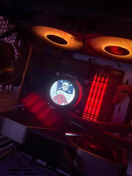 Gaming PC interior illuminated with red RGB lighting featuring fans, RAM sticks, and a circular display showing a skull-and-crossbones pirate logo with a pink creature.
