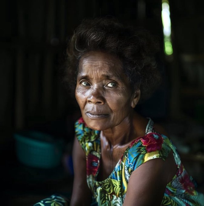 A portrait of an older woman with short dark hair wearing a colorful floral patterned top, sitting in a dimly lit interior with natural light filtering in.