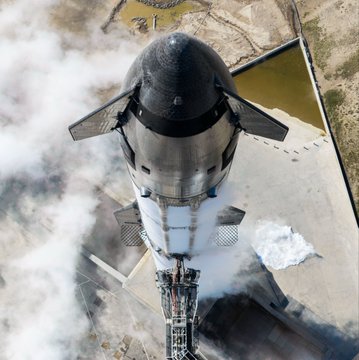 An aerial view looking down at a SpaceX Starship rocket on its launch pad, surrounded by steam and clouds.