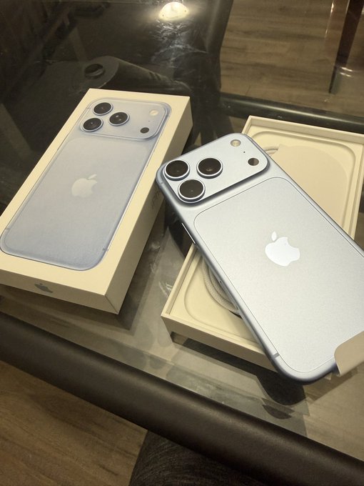 Two iPhone 13 Pro devices in Sierra Blue are displayed unboxed on a glass table, one still in its packaging showing the product image and the other resting on the box lid.