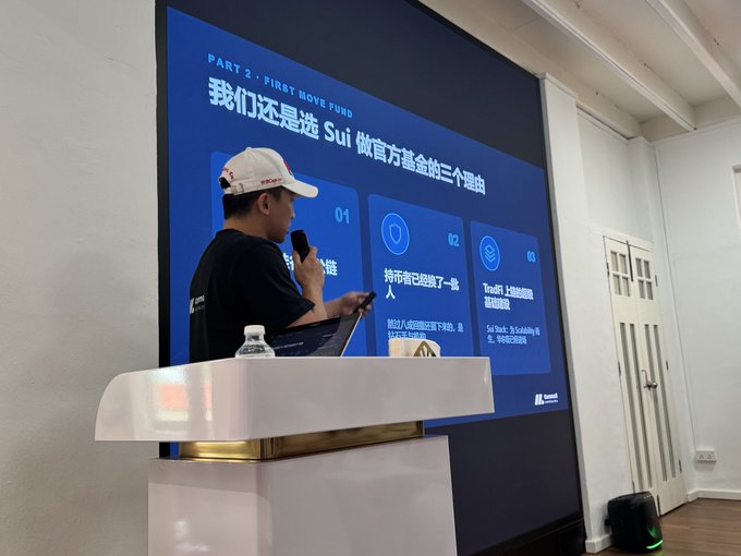 A speaker wearing a white cap stands at a podium holding a microphone and phone, presenting in front of a large screen displaying a Sui Foundation slide titled "PART 3 - FIRST MOVE FUND" with Chinese text explaining why to choose Sui as an official foundation, listing reasons like lower entry barriers and superior performance compared to other chains. The presentation relates to the post's mention of attending comma3vc's Morning Circle event, where the discussion appears focused on Sui's advantages as a blockchain investment opportunity.