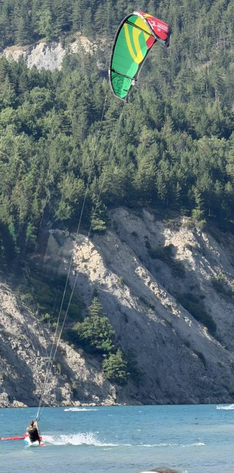A person kiteboarding on a body of water with a green and yellow kite, set against a backdrop of forested rocky cliffs.