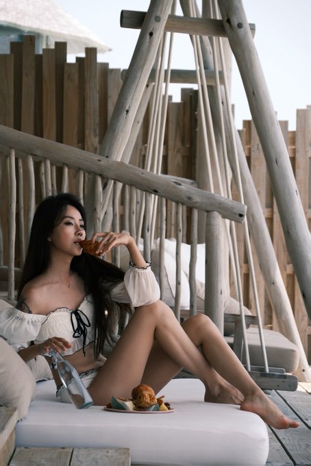A woman with long dark hair relaxes on a suspended net lounger at what appears to be a luxurious tropical resort, sipping a drink while a small plate of fruit rests nearby. The rustic wooden architecture and rope swing setting align with the post's reference to the high-end Soneva Jani resort in the Maldives.