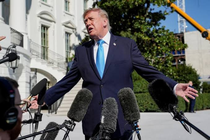 A photograph of Donald Trump speaking to reporters outside, wearing a dark suit with a bright blue tie and standing in front of multiple microphones.
