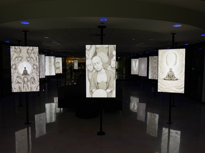 An art installation displays illuminated vertical panels featuring grayscale illustrations of meditating figures and abstract imagery in a dimly lit gallery space.