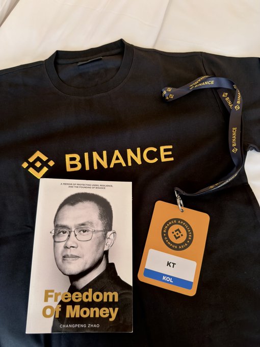 A black t-shirt with the gold 'BINANCE' logo and lanyard, accompanied by a book titled 'Freedom Of Money' featuring Changpeng Zhao (CZ).