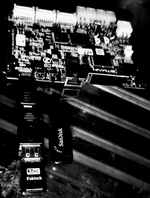 A black and white photograph shows computer hardware components, including a Bitmain-branded circuit board, a SanDisk Cruzer USB drive, and an AC adapter with the 'Foktech' brand.