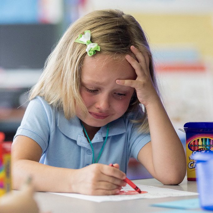 A young blonde girl with a green bow in her hair sits at a desk, frowning while concentrating on coloring or writing with a red marker, with a Crayola cup nearby.