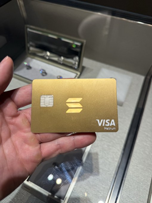 A hand holds a gold Solana Visa Platinum debit card in front of a display case containing jewelry.