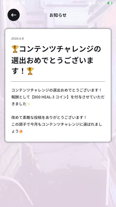 The image shows a phone screen with an app that has Japanese text displayed, possibly written in English as well. The text appears to be about a competition or achievement, and there are two gold trophies shown within the app. One trophy is located on the left side of the screen while the other one is positioned more towards the center.