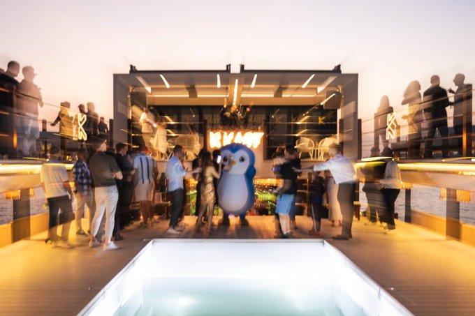 A crowd of people mingles on the deck of a yacht at dusk around a large penguin mascot, with the word "KAST" displayed on a screen behind them. The motion-blurred guests and central penguin character reinforce the post's playful "Spot the CEO" challenge, framing the CEO search as a lively networking event hosted by KAST.