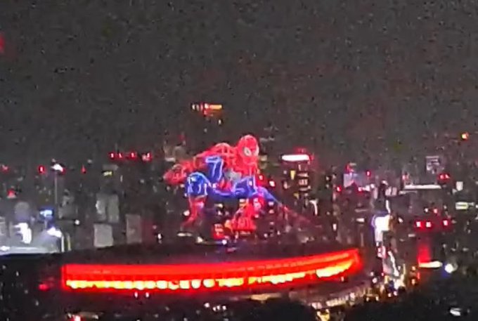 A nighttime cityscape shows a large illuminated Spider-Man figure displaying the number "7" on its chest, glowing in red and blue above a brightly lit red rooftop structure, seemingly connecting to the post's playful reference to the iconic Spider-Man theme song and the "7" symbol.