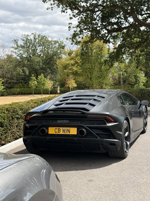 A matte black sports car (appearing to be a Lamborghini Huracán) is parked outdoors with a UK license plate reading "CB WIN," cleverly mirroring the "CB wins @coinbase maxxing" theme as a personalized vanity plate celebrating a win.