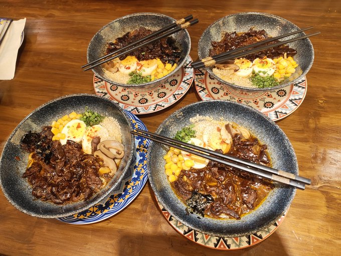 Four bowls of Asian-style braised meat rice with corn, egg, and mushrooms, each served with chopsticks on decorative patterned plates on a wooden table.
