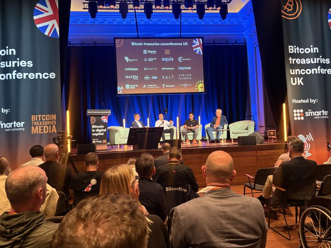 A group of people are sitting down, watching a presentation at an event with a large screen behind them displaying information about Bitcoin. The audience appears engaged as they listen to the speakers discussing the topic.