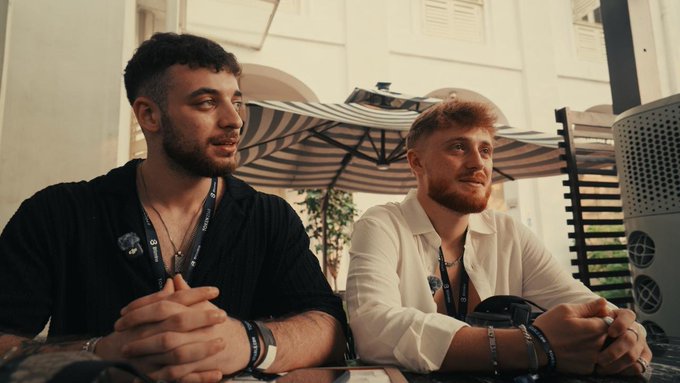 Two bearded young men wearing wristbands sit side by side at what appears to be an outdoor event, with a striped umbrella and white building in the background.