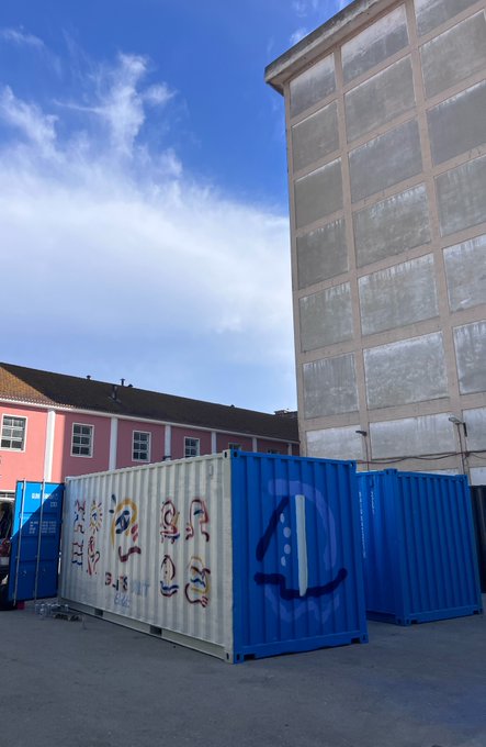 The image features a large blue shipping container with white lettering that reads "4" and "Lisbon". The container is situated outside of a building, possibly an office or warehouse. In front of the container, there are two smaller blue containers painted in white. A person can be seen standing near these containers, likely overseeing their placement or maintenance.
