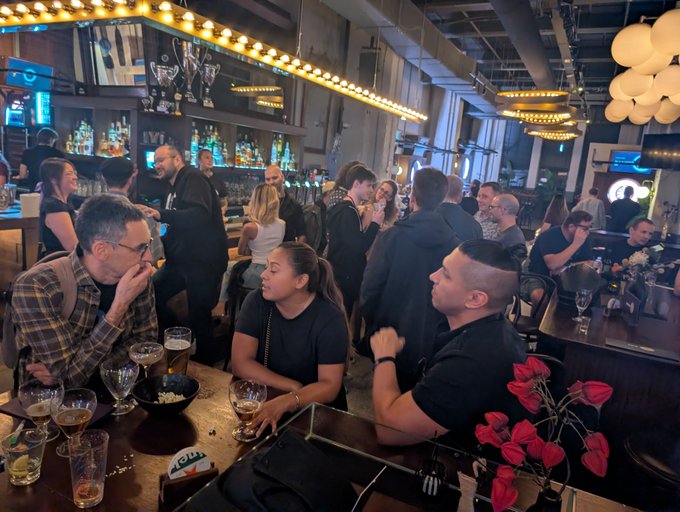 A crowded bar scene with patrons socializing at the counter, featuring warm lighting from string bulbs and modern spherical pendant lights.