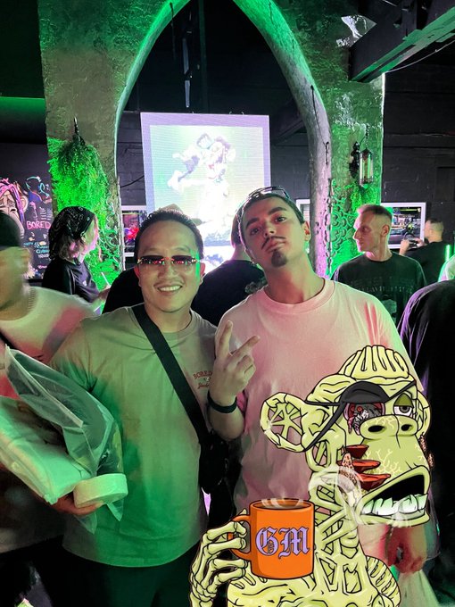 Two men pose together for a photo in a dimly lit venue with green lighting and arched stone walls, one holding a packaged item.
