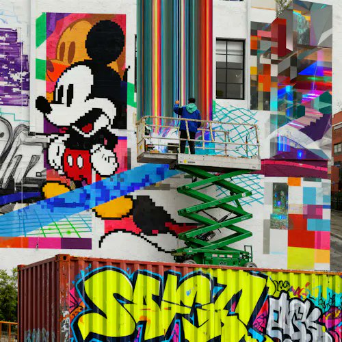 A large outdoor mural featuring a pixelated Mickey Mouse character with vibrant colors and geometric shapes, with a scissor lift in front.