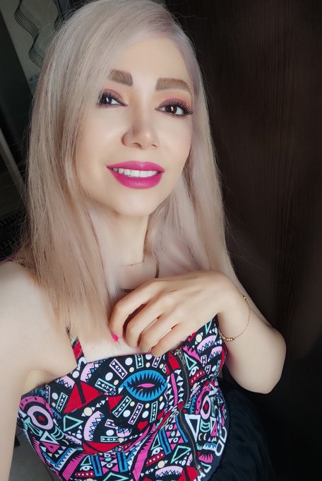 A selfie of a smiling woman with long blonde hair wearing pink lipstick and a colorful geometric-patterned top.