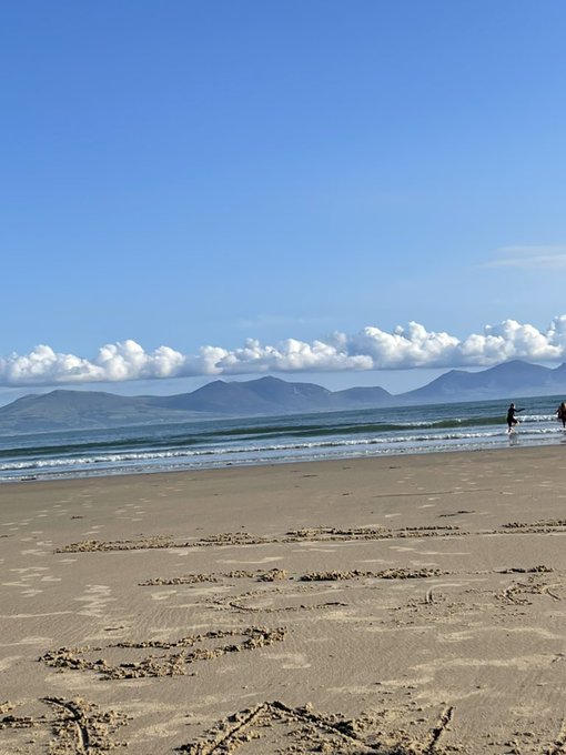 A scenic beach view featuring sandy shore with footprints in the foreground, gentle waves, distant mountains under a blue sky with scattered clouds, and a couple of people walking near the waterline.