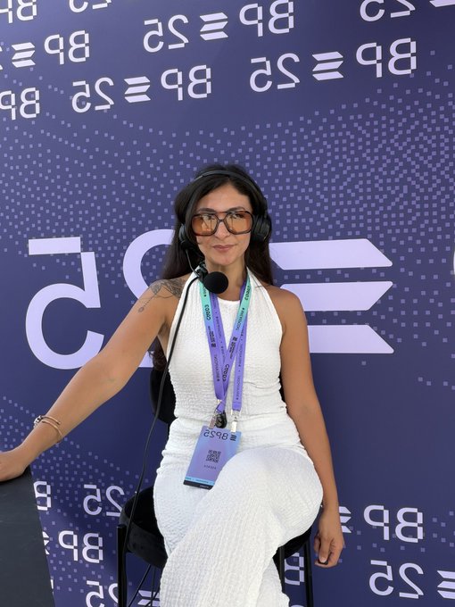 A woman wearing headphones, sunglasses, and a press badge (number 2596) sits in front of a blue backdrop featuring the "BTC25" logo, appearing to conduct interviews at a crypto conference. The image aligns with the post's message about her active role in covering the Solana community and blockchain events, supporting her nomination for an award.