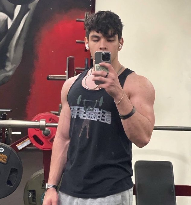 A muscular young man taking a mirror selfie in a gym, wearing a black tank top that reads "UNTRAINED", white wireless earbuds, and a black fitness watch, with weightlifting equipment visible behind him.