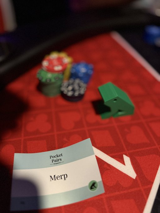 A close-up of a casino-style card game setup featuring stacks of colorful poker chips and a green chip holder on a red felt surface, with a card reading 'Pocket Pairs — Designers — Merp' in the foreground.