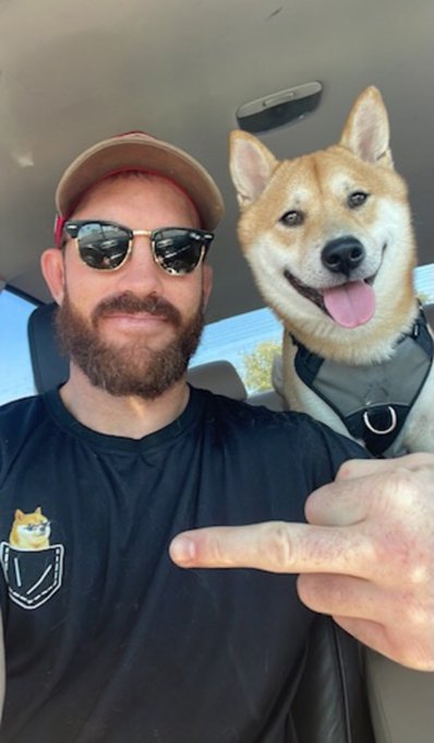A bearded man in sunglasses and a cap smiles in a car beside a happy Shiba Inu wearing a harness, while he points at his shirt featuring a matching Shiba Inu logo. The post’s “Can we get a GM?” seems to be a playful call for good morning, paired with their cheerful morning selfie.