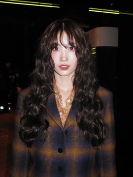 A woman with long wavy dark hair and bangs wears a plaid blazer, standing in what appears to be an indoor venue.