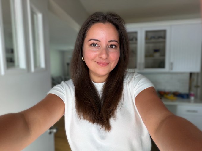 A woman with long brown hair wearing a white t-shirt takes a selfie in what looks like a kitchen or dining area.