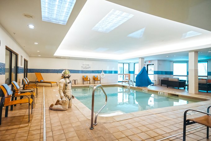 An astronaut in a white spacesuit kneels by the edge of an indoor hotel pool, with orange lounge chairs lining the tiled deck and a sign on the wall reading 'NO DIVING'.