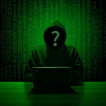 A hooded, faceless figure with a question mark sits behind a laptop, surrounded by cascading green code reminiscent of "The Matrix," visually evoking the mystery and digital theft behind the Bitcoin hardware wallet breach that stole $38 million from 500 supposedly offline wallets in just 25 minutes.