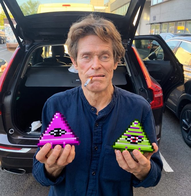 A man with messy hair and a cigarette in his mouth stands in front of an open SUV trunk, holding two pixel-art triangular objects with eyes—one pink/magenta and one green.