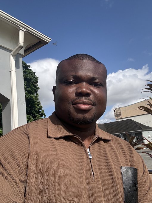 Selfie photo of a man in a brown zip-up shirt taken outdoors with houses and a partly cloudy sky in the background.