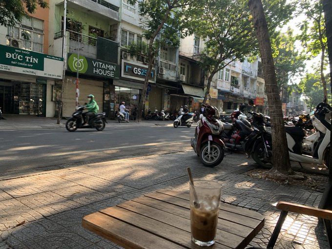 A street view of a Vietnamese urban area featuring storefronts including 'Đức Tín', 'Vitamin House', and 'FGS', with motorbikes parked along the sidewalk and an iced coffee on a wooden table in the foreground.