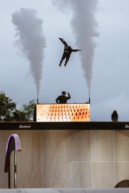 Action shot of a skateboarder mid-air performing a trick above a halfpipe ramp, with two smoke plumes rising behind them on a SkateLite-branded stage.