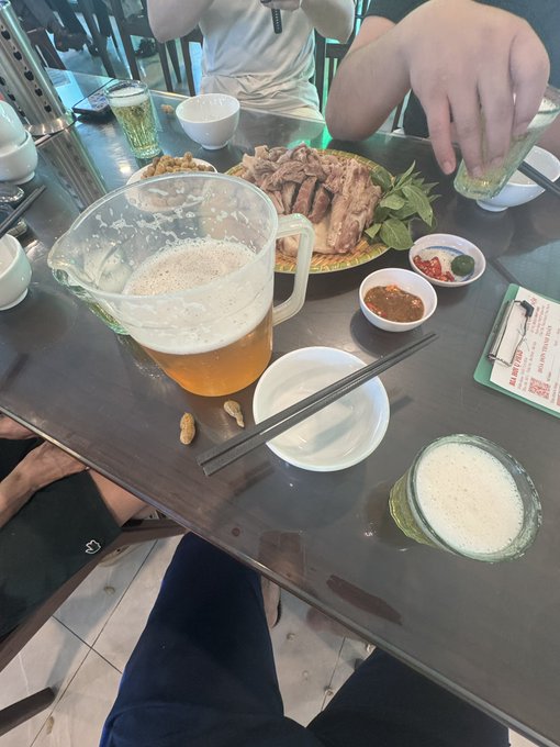 A first-person perspective of a meal at a restaurant table with a metal surface, showing a pitcher of beer, a glass of beer, a plate of sliced meat with mint leaves, dipping sauces, chopsticks, and peanuts scattered on the table.