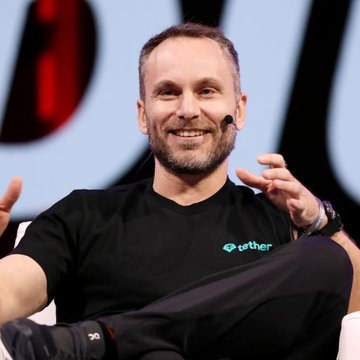 Tether CEO Paolo Ardoino, wearing a branded black t-shirt with the "tether" logo, speaks at an event with a headset microphone while gesturing during a panel discussion. The image aligns with the post highlighting his critique of excessive AI infrastructure spending by major tech companies.