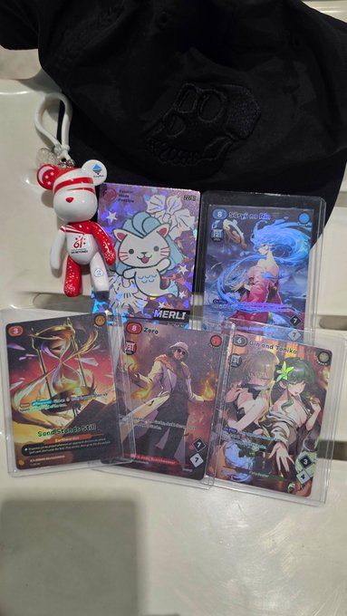 A collection of trading cards featuring anime-style art with character names 'MERLI', 'Zero', and 'Rena Yuko', alongside a small red and white plush toy and a black bag in the background.