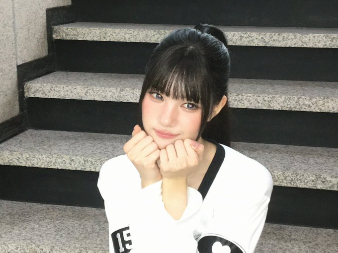 A young woman with dark hair and bangs sits on stone stairs, resting her chin on her hands and smiling at the camera while wearing a white shirt with a black strap.