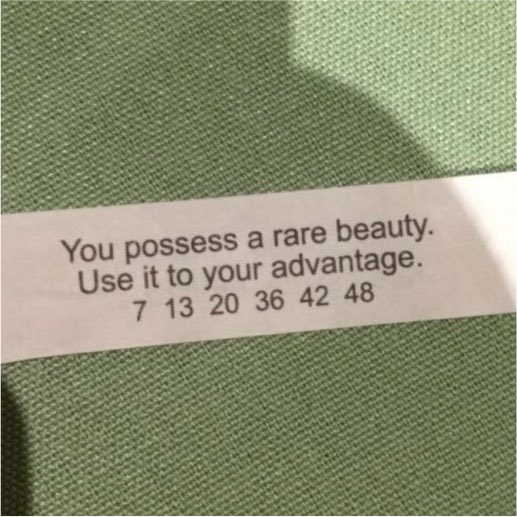 A close-up photo of a fortune cookie slip resting on green fabric, reading 'You possess a rare beauty. Use it to your advantage. 7 13 20 36 42 48'.