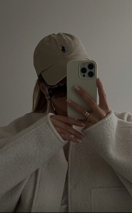A mirror selfie showing a person wearing a beige cap with a dark logo, tortoiseshell sunglasses, and a light gray coat. They are holding a smartphone with a light-colored case that covers their face, with manicured nails and a gold ring visible.