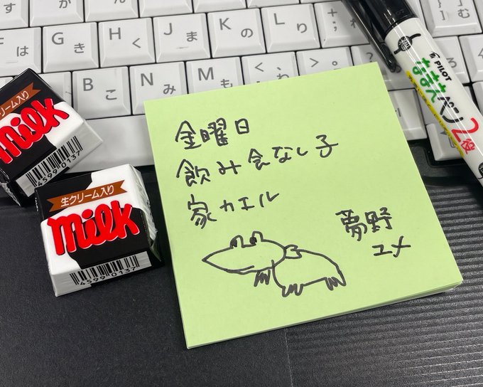 A green sticky note with Japanese handwriting reads '金曜日飲み会ない子 かれにし 南野ユキ' with a small crocodile drawing, placed beside two 'milk' cream candies, a Japanese keyboard, and a Pilot marker.