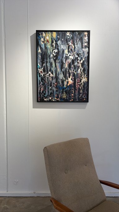 A framed abstract painting with dark blues, blacks, and earth tones hanging on a white wall above a beige mid-century chair.
