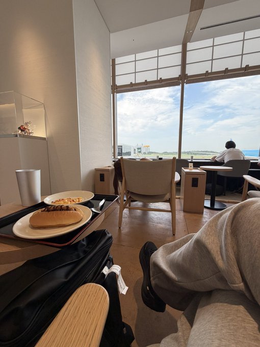 A view from a lounge area looking toward large windows, showing a tray with food in the foreground and a person relaxing with a view of the tarmac outside.