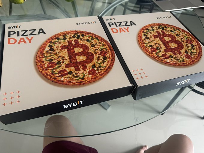 The image shows a table with two pizza boxes placed on top of it. The boxes are black and white, and they contain pizzas. The text "Bybit Pizza Day" can be seen on one of the boxes, indicating that the pizzas were ordered from Bybit. The price for these pizzas is 10,000 BTC, which is a significant amount in today's currency exchange rates. However, due to low liquidity and increased interest in US stocks and gold, the value of the Bitcoins has dropped significantly, making the pizzas now worth only $746.5M USD.